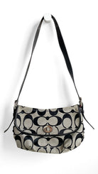 A Coach shoulder bag in the signature monogram canvas, featuring black and beige pattern with a black leather strap and a gold-tone turn-lock closure, hanging against a plain white wall.