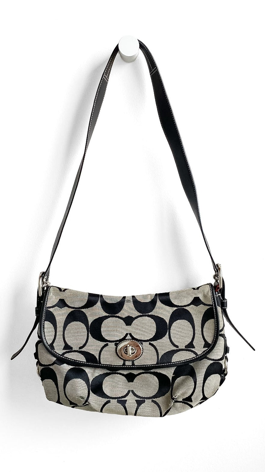 A Coach shoulder bag in the signature monogram canvas, featuring black and beige pattern with a black leather strap and a gold-tone turn-lock closure, hanging against a plain white wall.