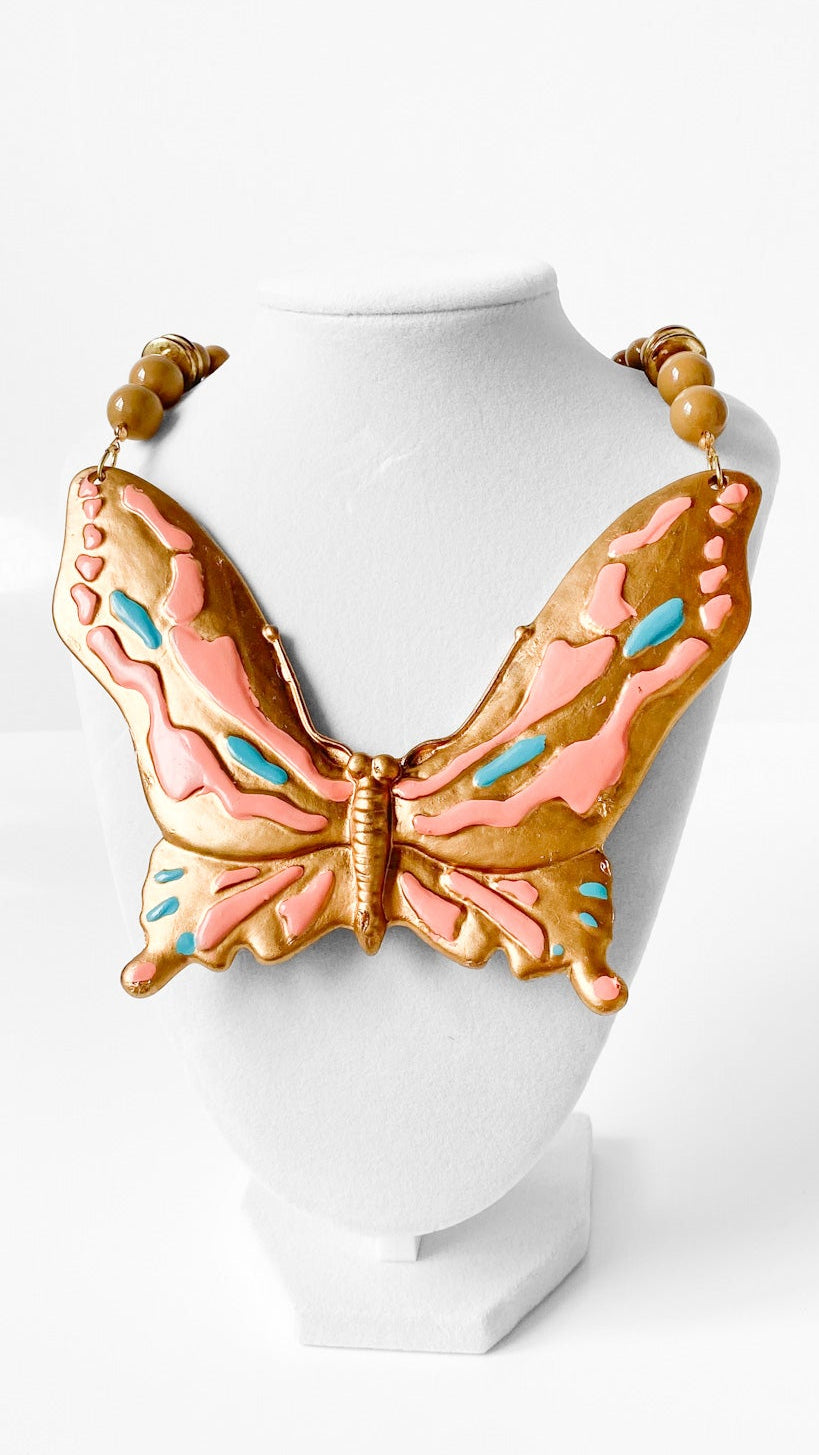 A butterfly-shaped statement necklace displayed on a white mannequin bust against a clean white background. The necklace features a large central butterfly with pink and orange wings, accented with light blue details, and a gold-tone body.