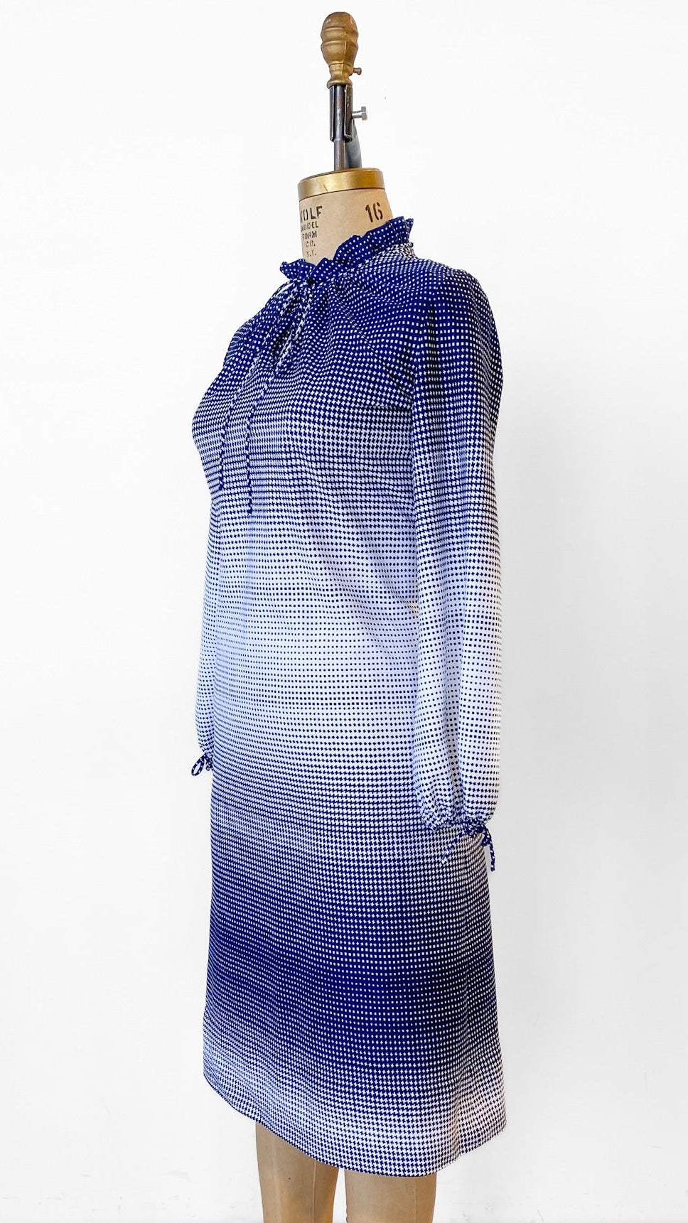 Mannequin wearing a blue and white checkered dress on a wooden floor with a white wall background.