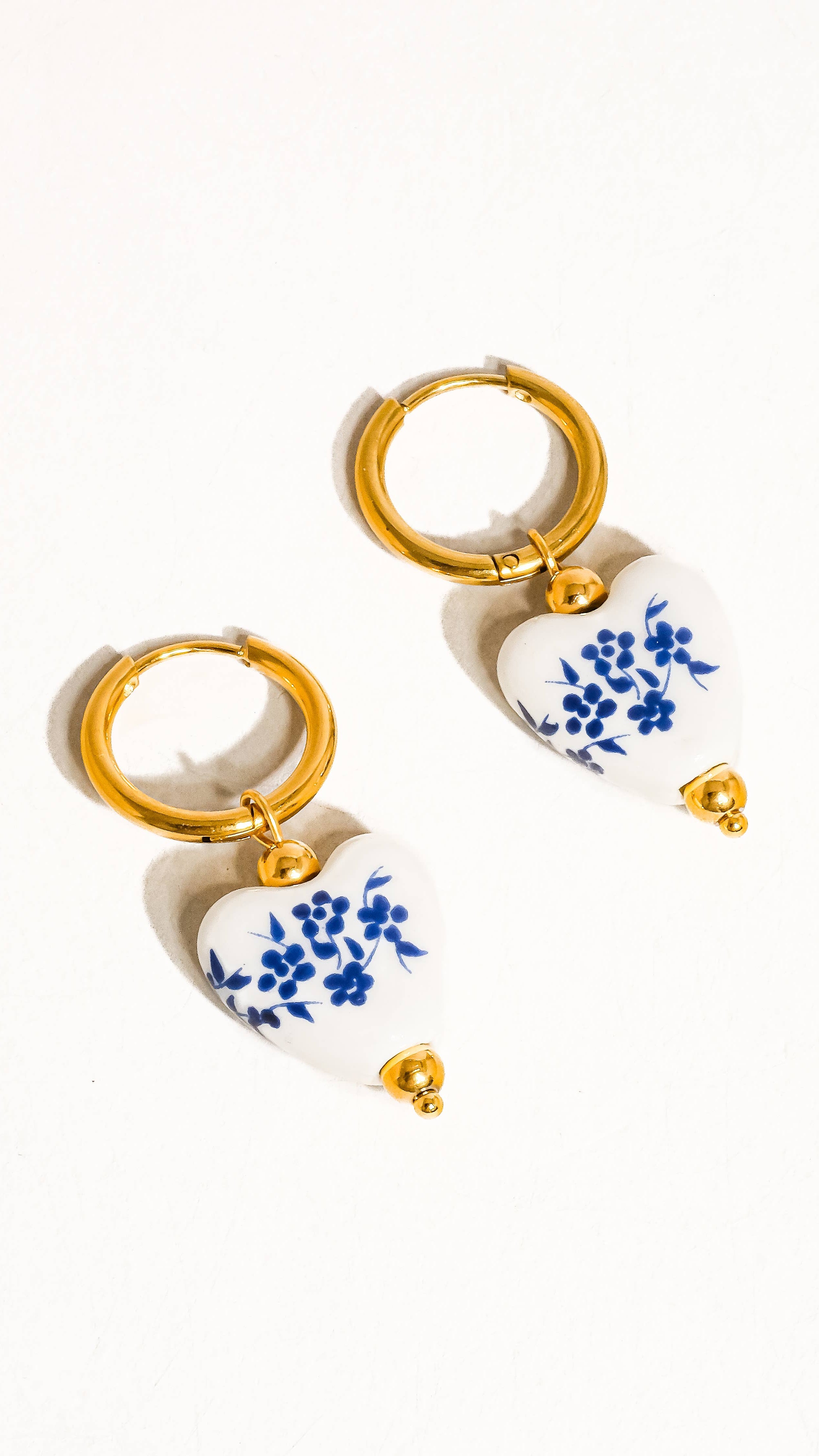 A pair of gold hoop earrings with ceramic floral heart pendants in blue and white.