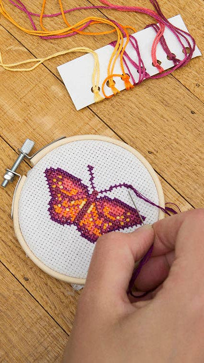 Person stitching a butterfly design on a cross-stitch hoop with colorful yarn on a wooden surface.